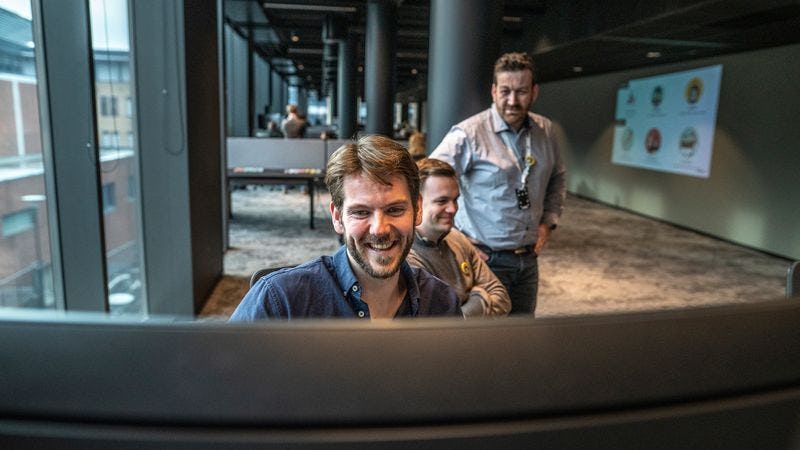Fornøyd Inmeta-konsulent foran stor PC skjerm i kontor på Nydalen med to kolleger bak seg
